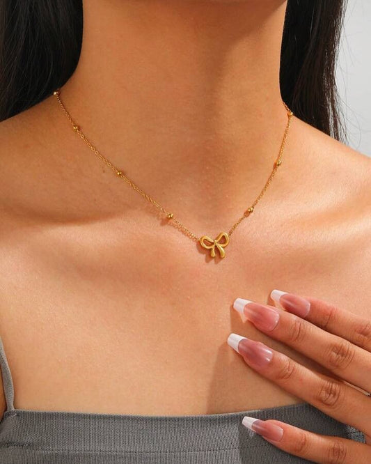 Gold necklace with a bow pendant worn by a person, with a hand touching the chest.