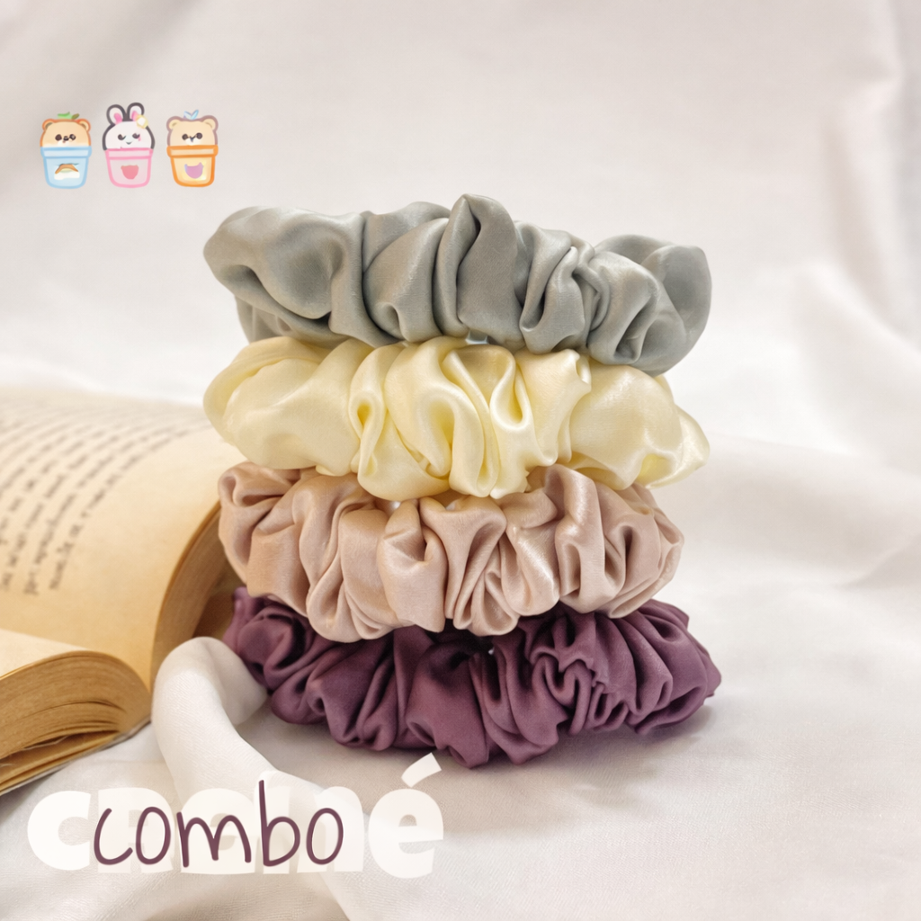 “Stack of four satin scrunchies in muted grey, soft yellow, beige, and deep plum, arranged on white fabric beside an open book, with small cute sticker icons adding a playful aesthetic.”